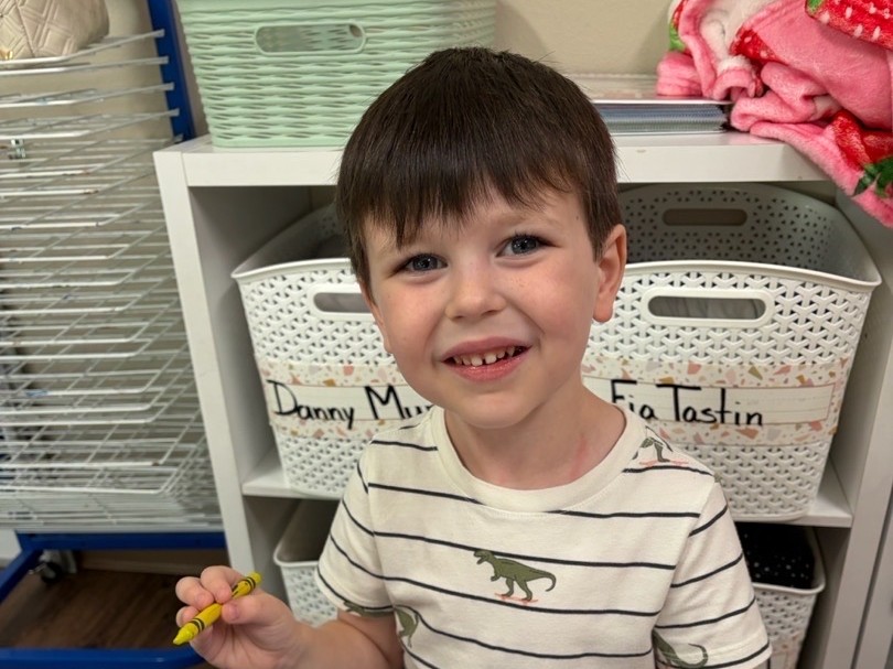 A preschool boy holding a yellow crayon beside his workbook at a classroom table.