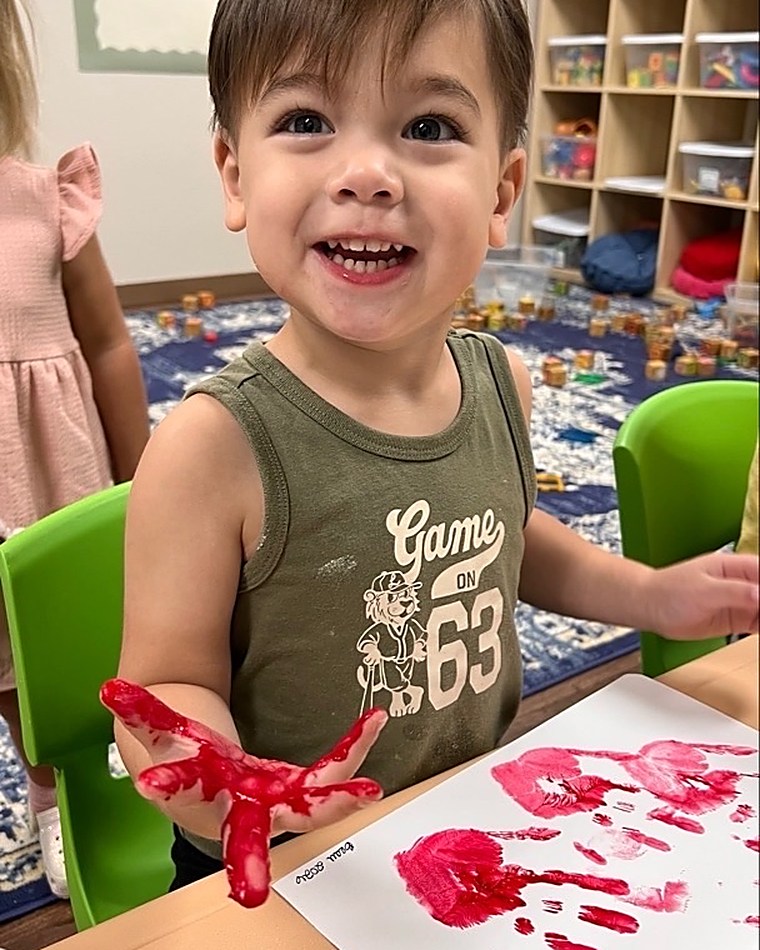 A smiling toddler holding up a paint-covered hand beside the artwork he made.