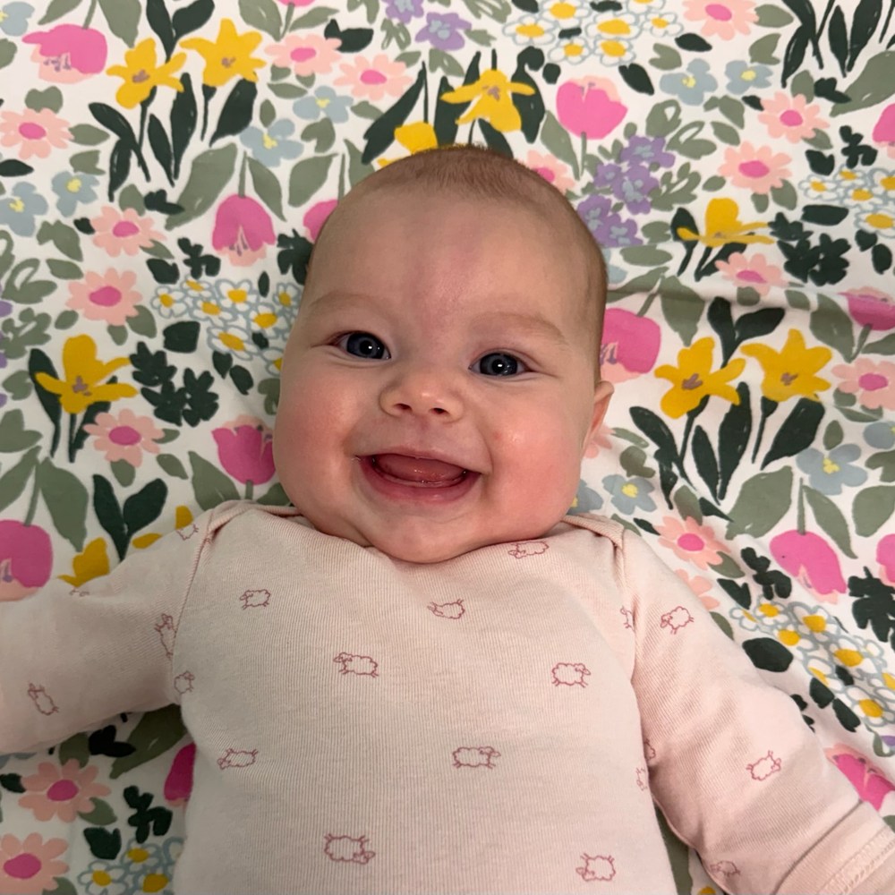 A smiling infant lying on a floral mat, looking up toward the camera.