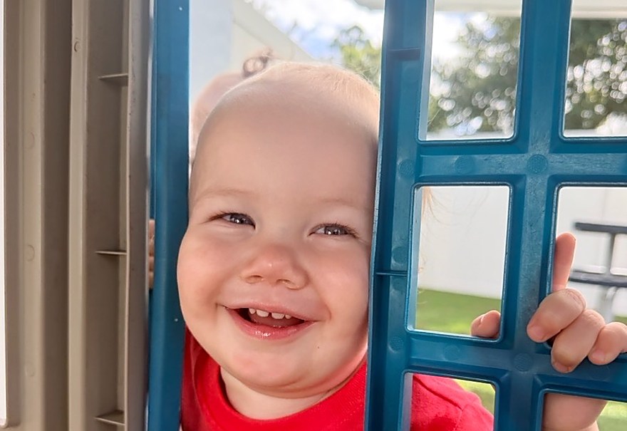 A smiling toddler peeking out of the window of an outdoor playhouse.