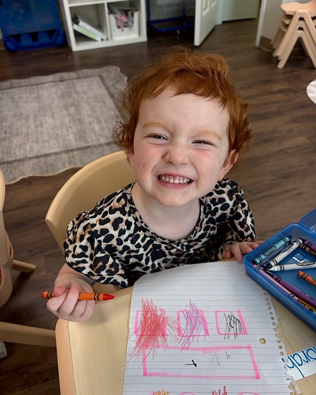 A smiling young child with curly hair colouring with a crayon at a classroom table.