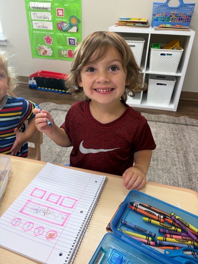 A preschool-age child painting at a classroom art table alongside other children.