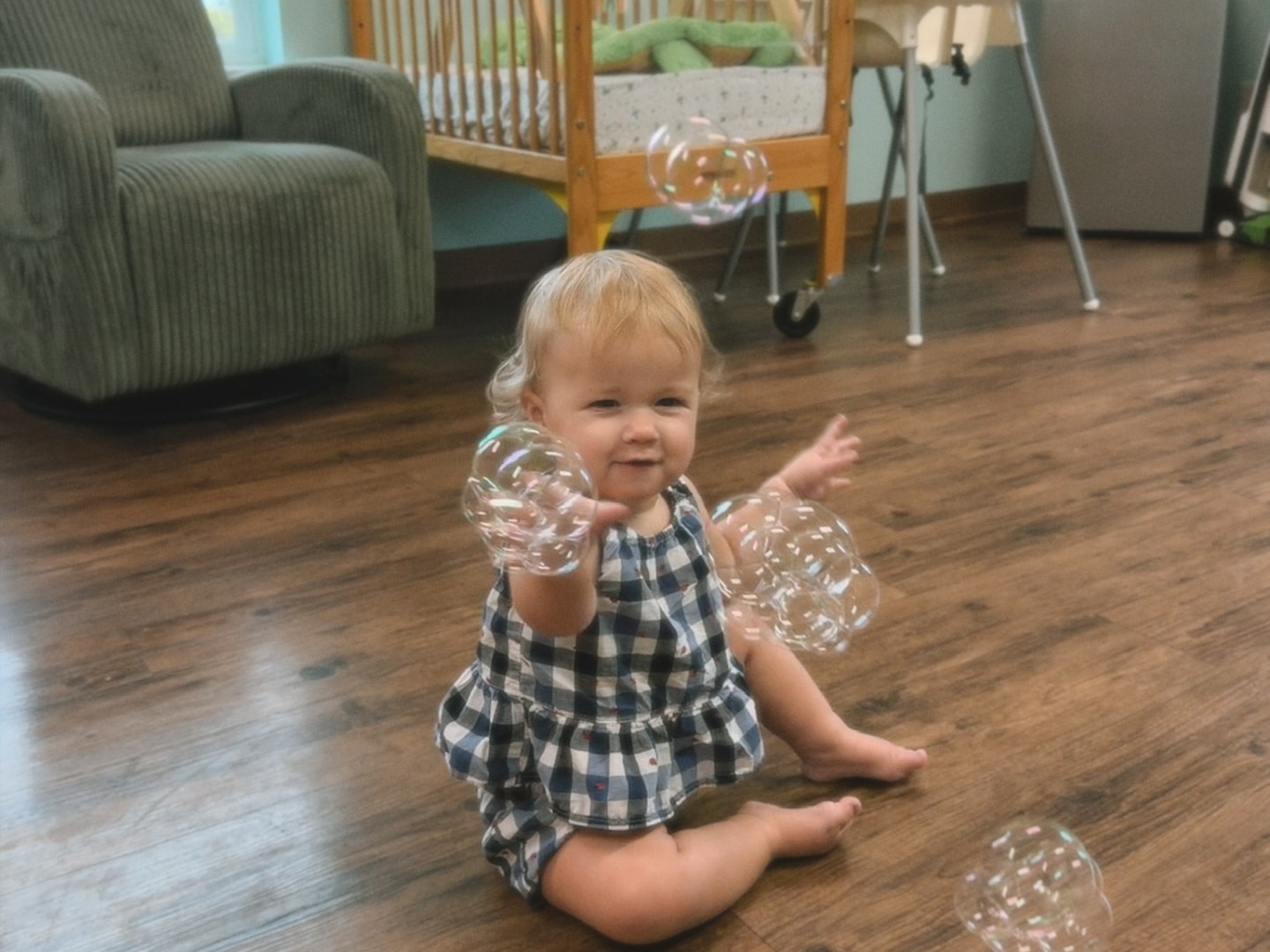 A smiling baby girl in a gingham romper sitting on a classroom floor, reaching up toward floating soap bubbles.