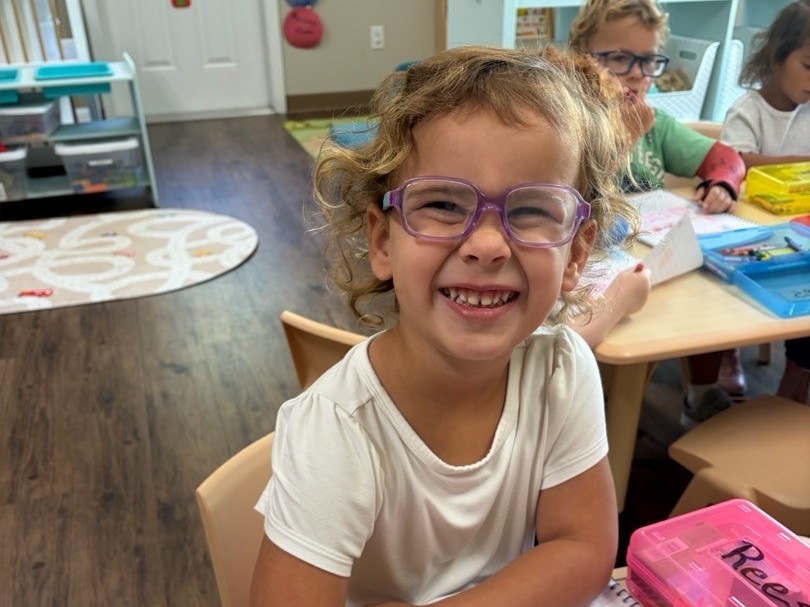 A smiling preschooler in purple glasses sits at a classroom table drawing with crayons on a workbook page.