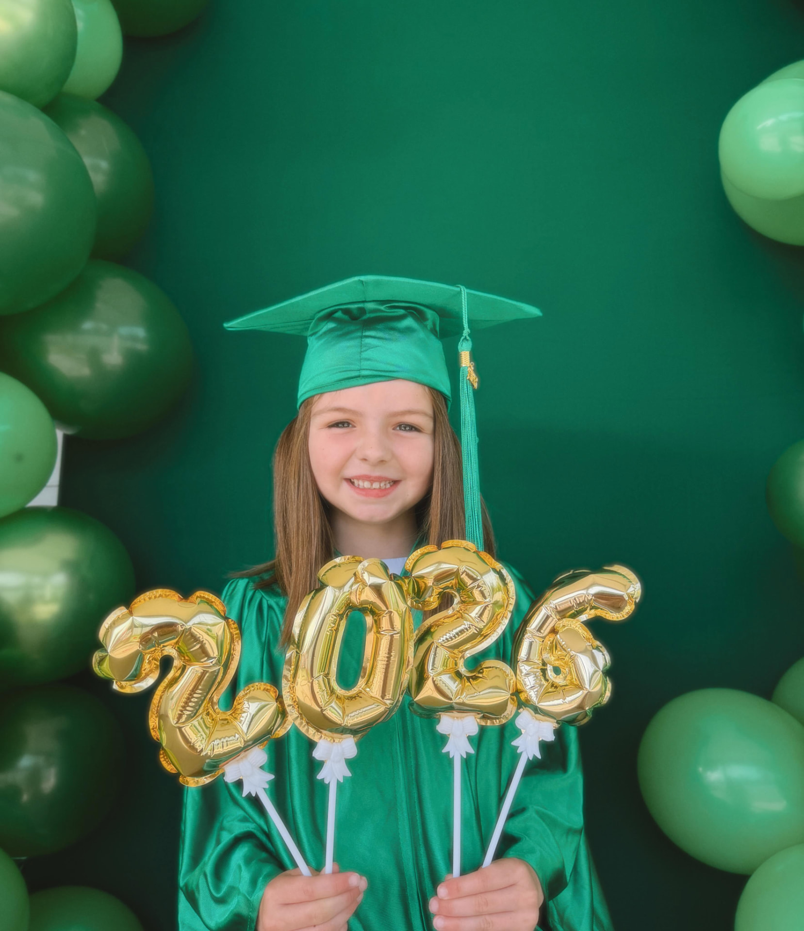 A smiling preschool graduate in a green cap and gown holds golden 2025 balloons.