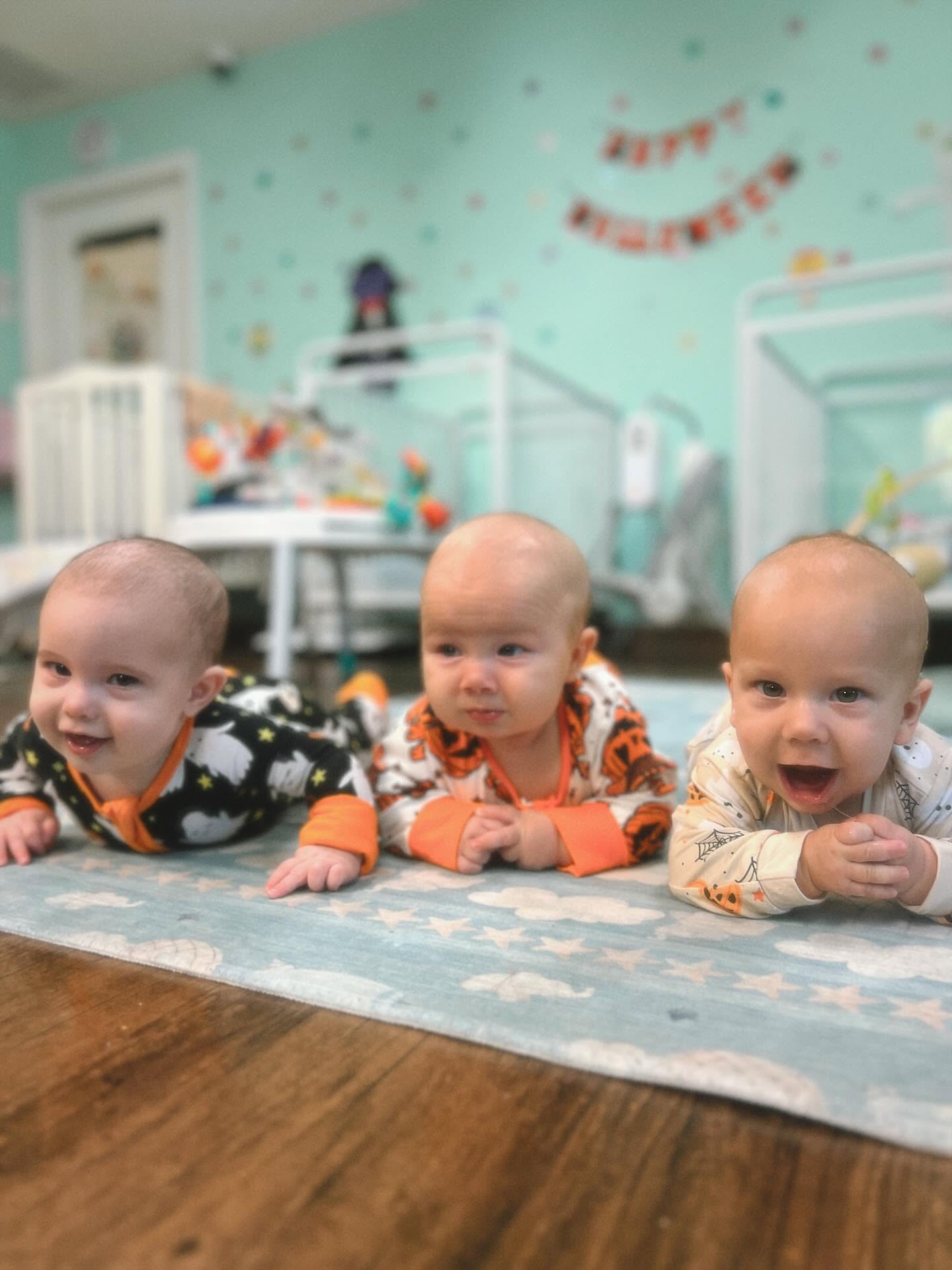 Three babies in costume pajamas smile during tummy time on a soft star-print mat in the Tadpole Prep infant room.