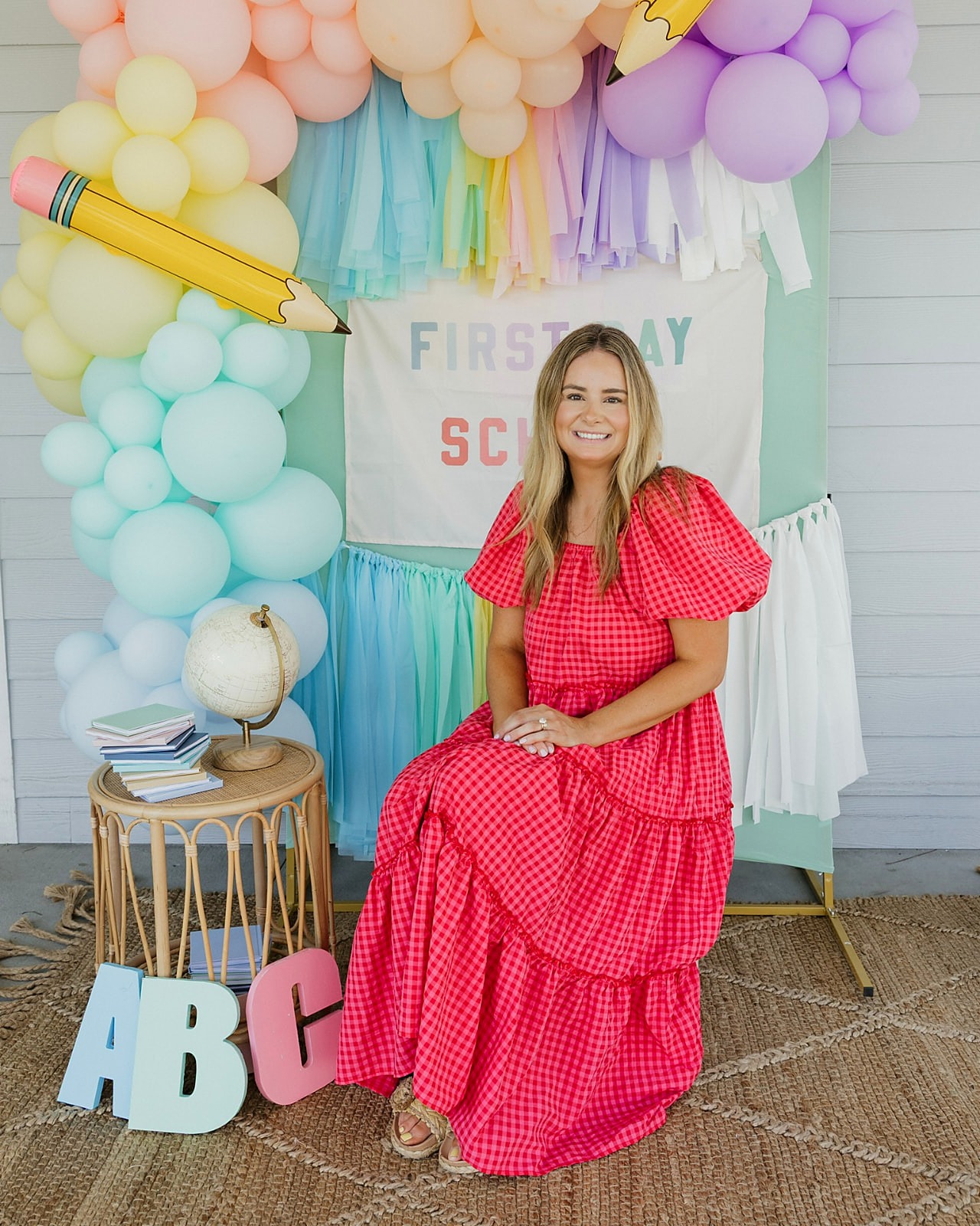 A smiling woman in a red gingham dress sits in front of a back-to-school backdrop with a pastel balloon arch, oversized pencils and ABC blocks.