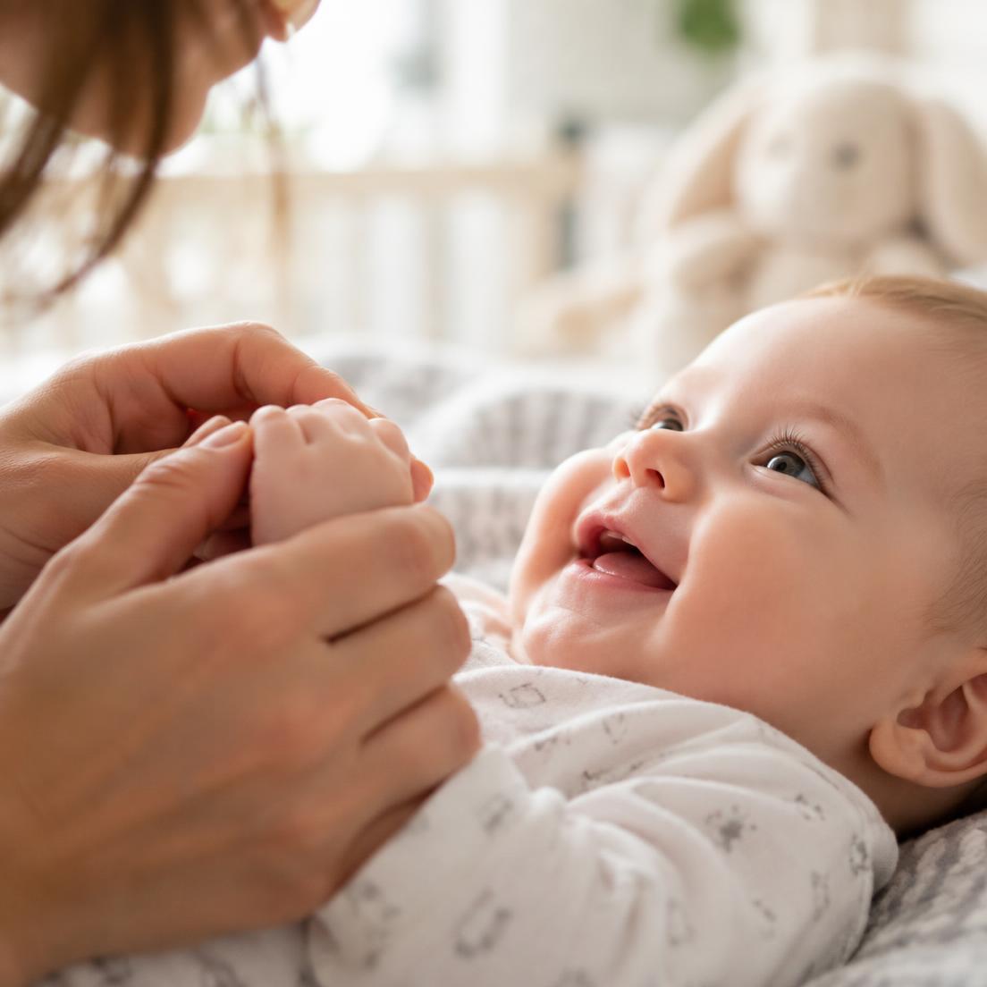 A caregiver gently holds a smiling baby's hand in a soft, bright nursery.